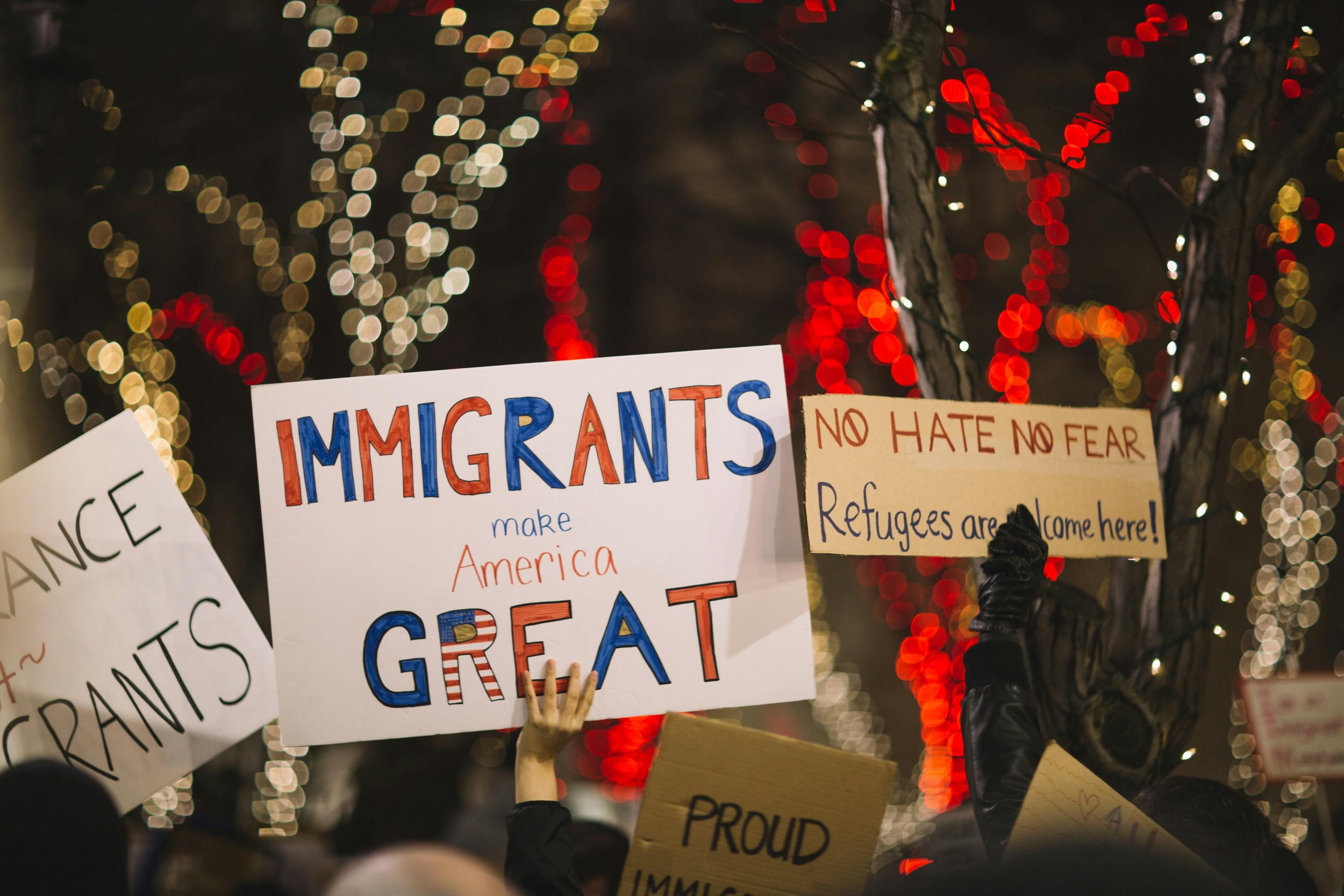 Group of protestors holding up signs protesting American immigration and border policies. Sign in middle says "Immigrants make America GREAT" in red and blue letters, and sign on the right says "NO HATE NO FEAR" on the first line and "Refugees are welcome here" on the second line. 