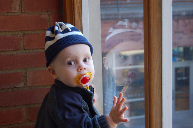 A young baby with a soother in their mouth, stares at the camera as they grasp the window that displays their reflection