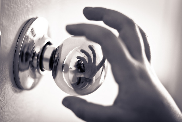 Black and white image of a hand reaching to grab a door handle. The door handle has a reflection of this in action.