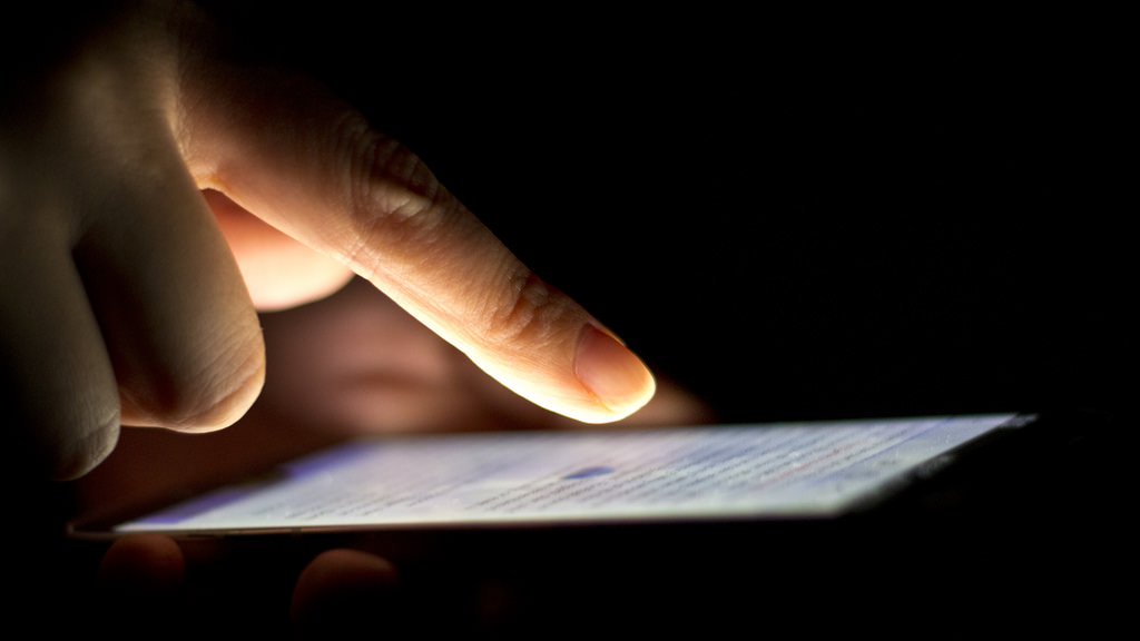 In a dark background, a human finger touches the glowing screen from a smartphone.