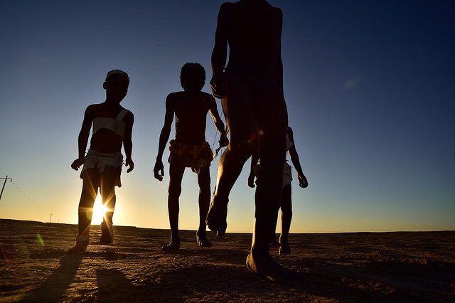 Silhouette of three figures walking with bare feet across the ground. In the distance is the sun rise.