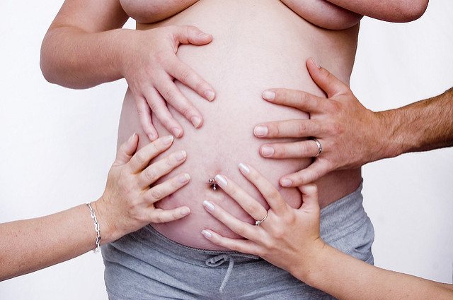 A bare torso of a pregnant woman is centrally touched by four different hands.
