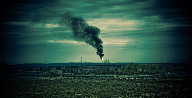 Far away landscape image of a factory, with smoke pollution. The image is filtered by a green and blue tint.