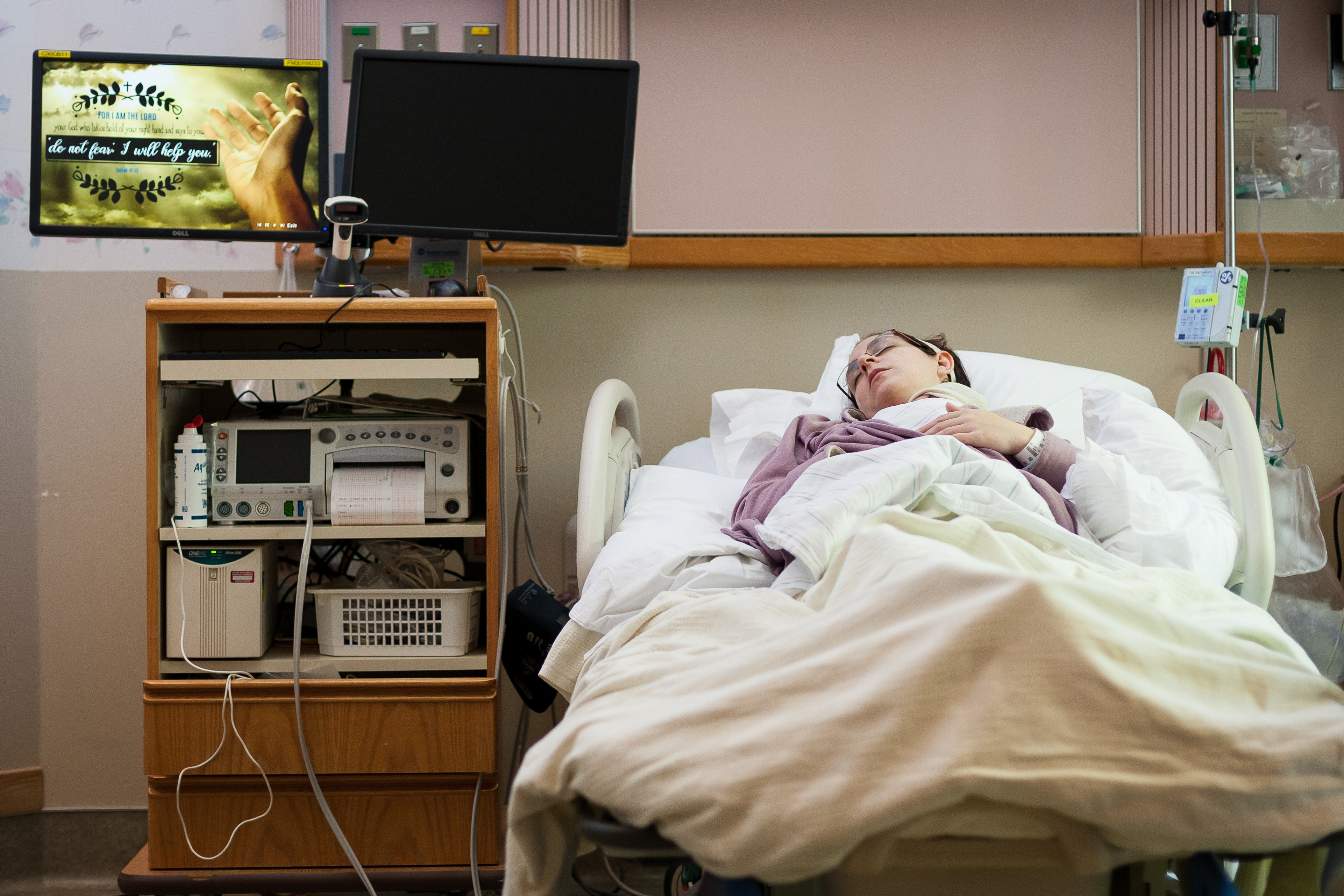 Female hospital patient lays in bed appearing asleep. Beside her is a TV.