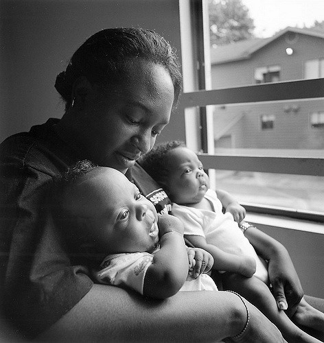 A black mother holds two babies in her arms. the babies look curiously out the nearby window.