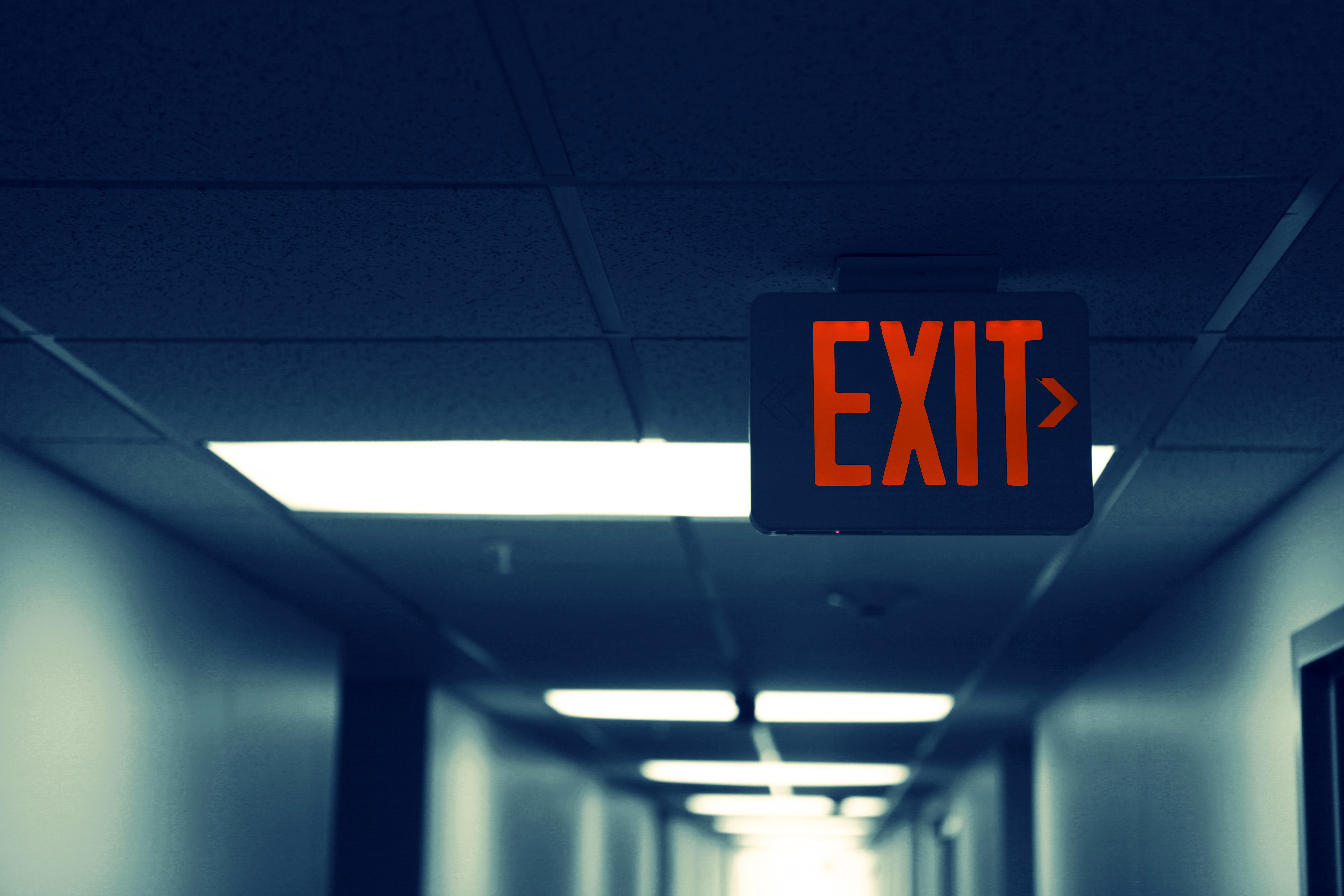 A red-glowing exit sign hangs above a hall way's ceiling.
