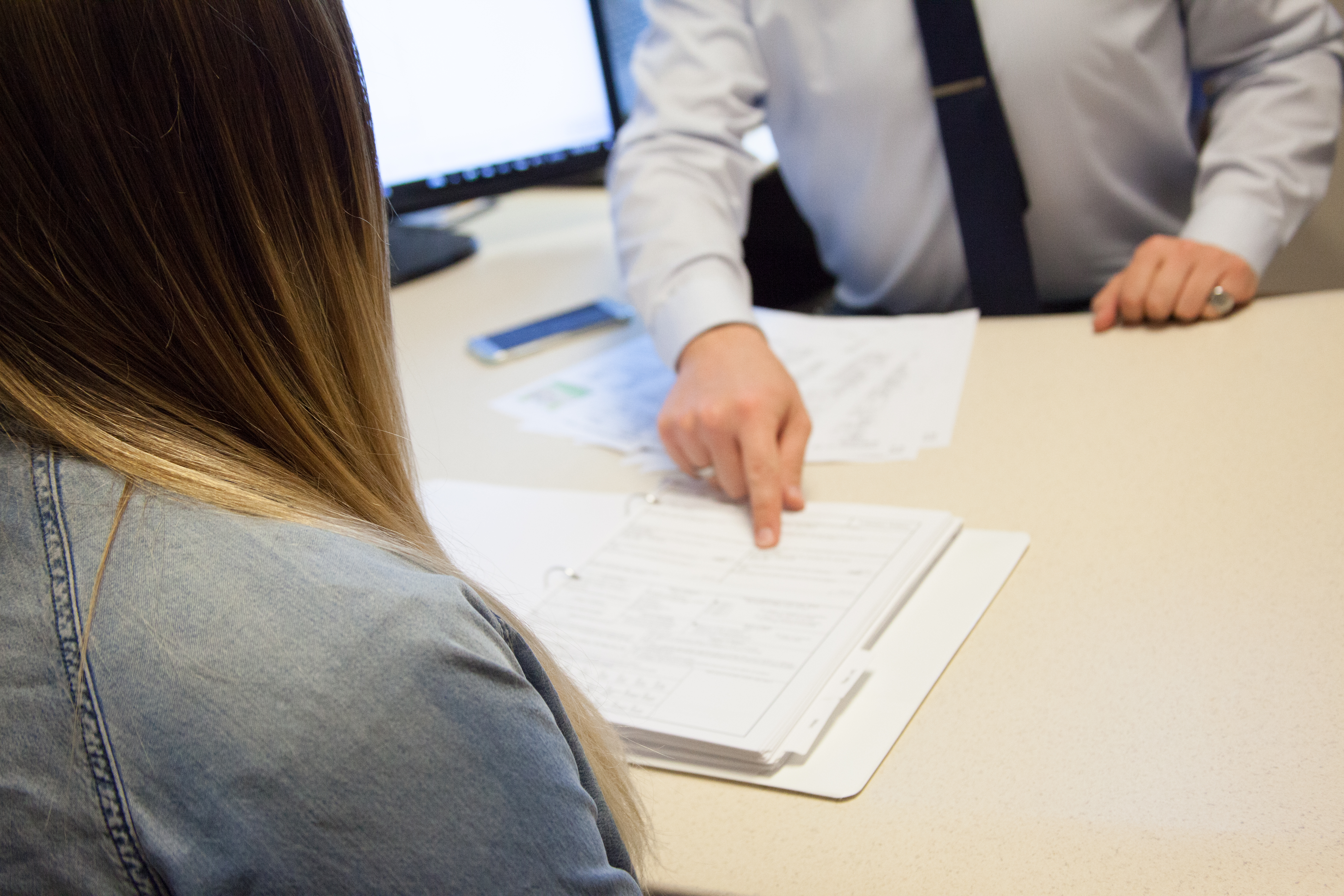 A woman's back is turned away from the camera. In front of her, a man in a suit points at a piece of paper within a binder on his desk.