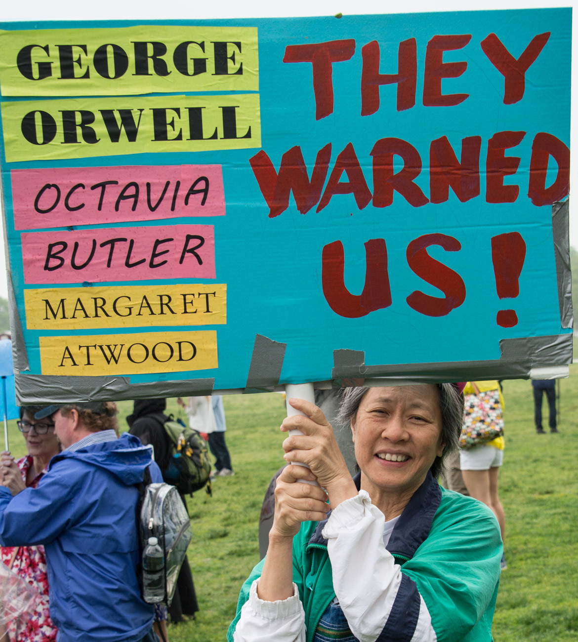 A woman of color holds a demonstration sign reading, "George Orwell, Octavia Butler, Margaret Atwood--They warned us!"
