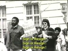 Black and white screenshot of a scene in the documentary The Power of 504, which highlights a victory in disability justice (reflected in Section 504 of the 1973 Rehabilitation Act). Protesters advocate for the civil rights of people with disabilities. Some hold signs stating "Our biggest handicap is [HEW Secretary Joseph] Califano!" Caption reads "Sign or resign!"