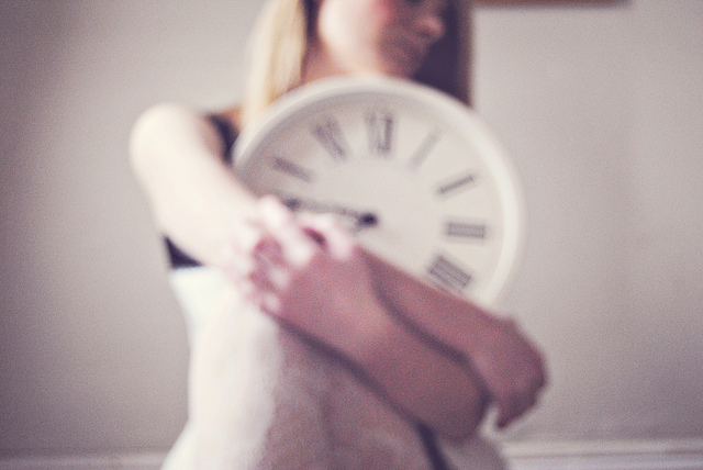 A feminine figure is sitting, holding a clock between her arms. The figure herself is out of focus.