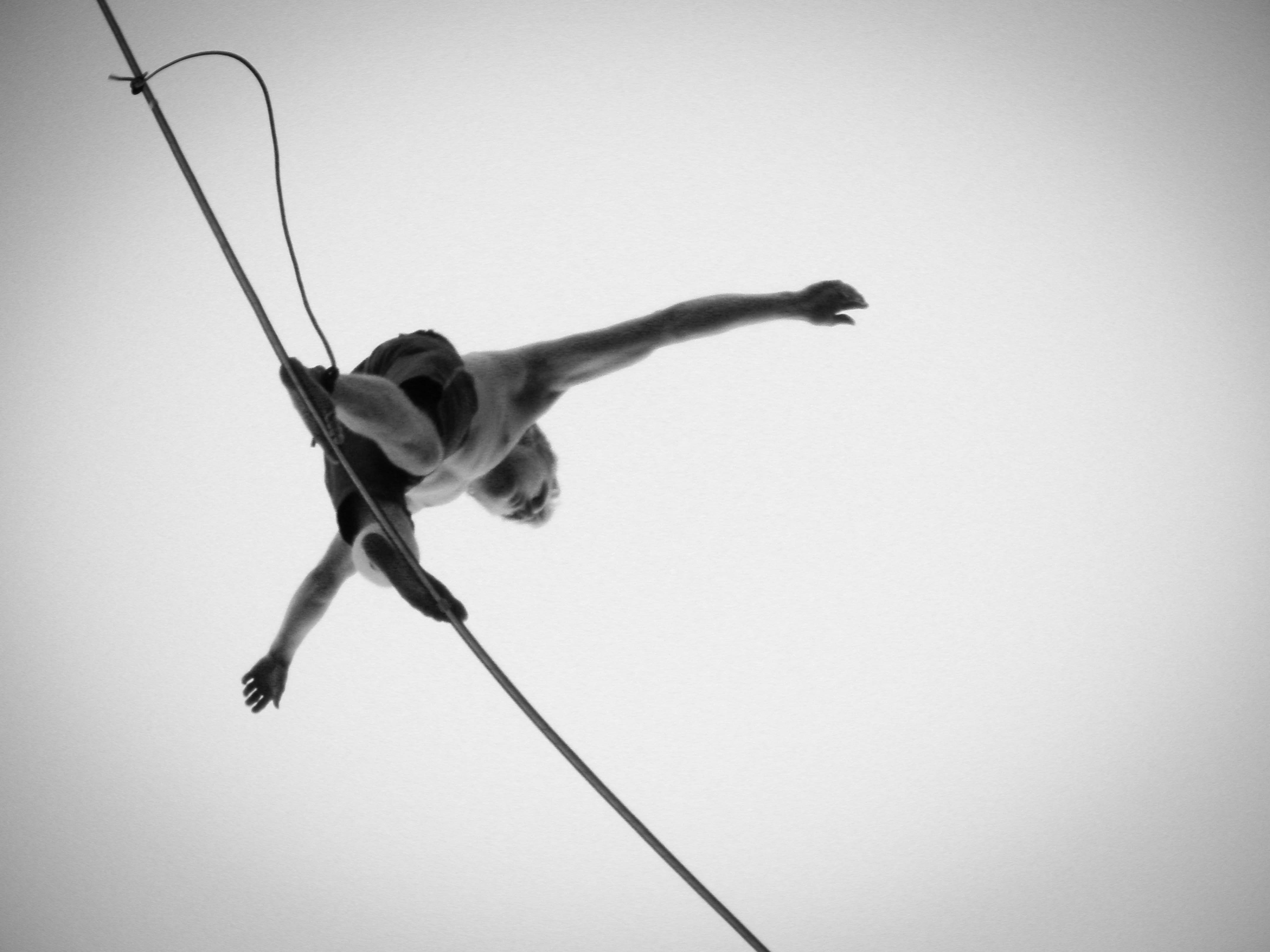 Black and white photo, in bug's eye view, looking up at a person balancing on a tightrope.