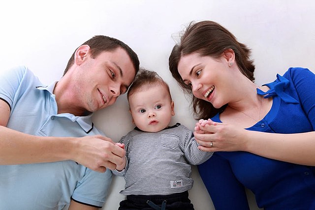A mother and father lie down with their baby in between them. They smile and look at the baby while holding its hands. The baby looks off into the distance.