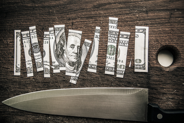 A one-hundred dollar bill is cut into pieces on a cutting board. Below the bill is a kitchen knife.