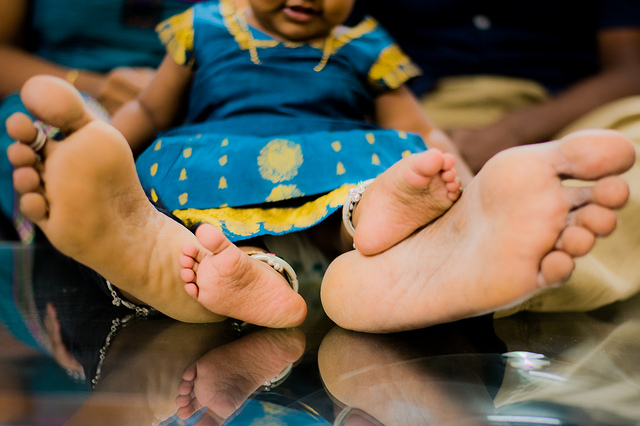 Adult feet and baby feet are next to each other. They are positioned as if the adult is sitting down and the baby is between their legs. They both wear bright blue clothing.