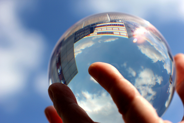 Human hands hold up a crystal ball to the sky, reflecting the blue sky with clouds and city buildings.