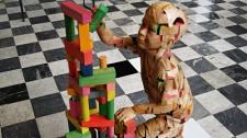 A wooden-like figure of a child builds a colorful tower of blocks. The child sits in a background surrounded by white and black checkered flooring.