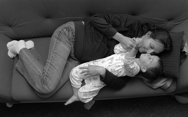 Grayscale image of an adult woman laying on a couch, with a toddler nestled in her arms. Both she and the toddler look affectionately at each other.