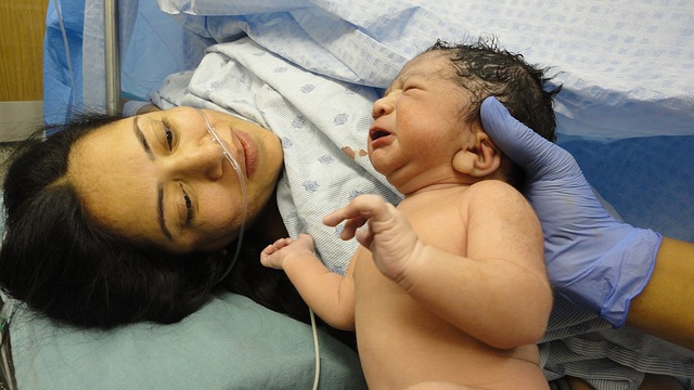 A young woman of color with an intravenous line is dressed in a hospital gown, and lays on the hospital bed, looking over to a newborn child that is supported by a gloved hand against the back of their head.