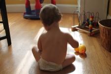 Silhouette of a baby. The baby's back is turned away from the camera, facing a window with sunlight. A rubber duck and several other toys are placed on the baby's side.