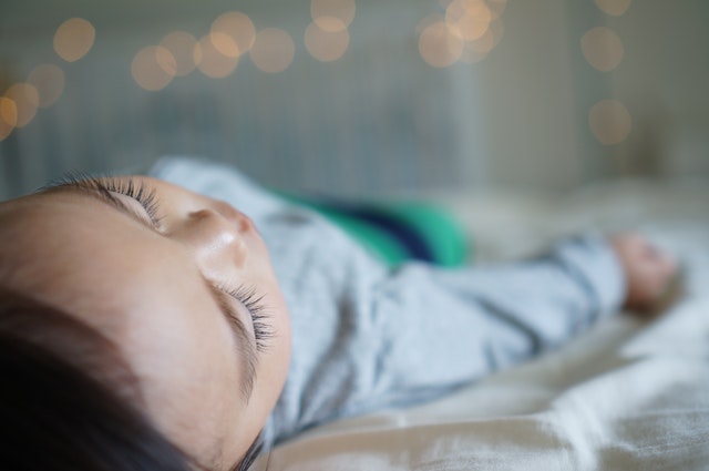 A close up photograph of a male Japanese baby lying down on a bed in pajamas with eyes closed.