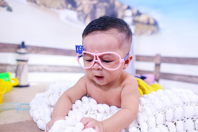 A baby plays with a blanket that they are laying on. The baby is wearing pink glasses frames without lenses, with the words "Kiss."