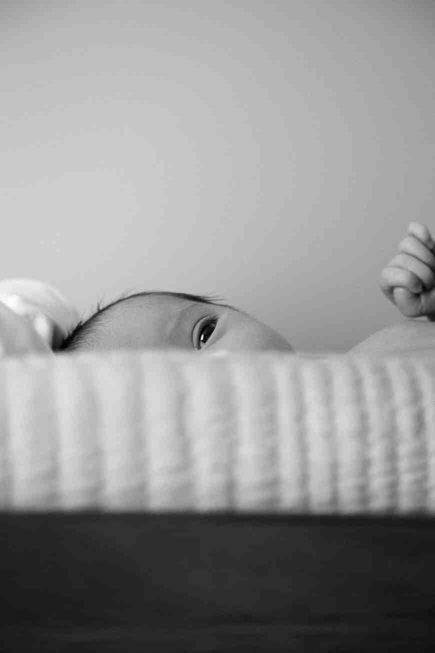 a black and white photo of a striped bed with a baby peaking over the edge. The left side of their face is visible.