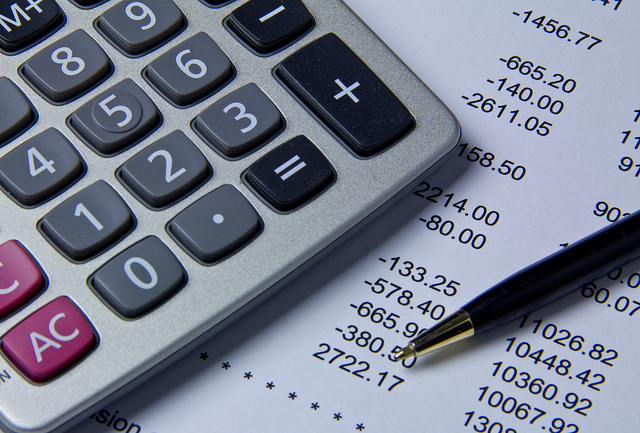A calculator rests on top of an sheet of paper with numbers that signify expenses. A pen rests nearby.