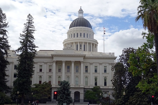 Landscape photo of the CA Capitol Building, against a blue sky and surrounded by trees,\.