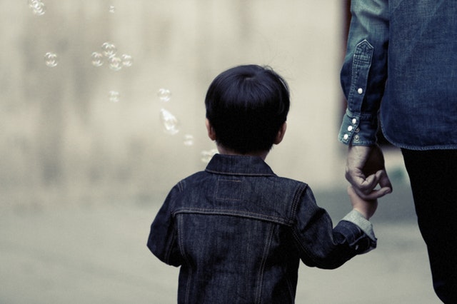The back of a child walking with an adult who is holding their hand. Surrounding the child are bubbles in the wind.
