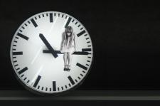 Black and white image. A woman sits peering down, on the long hand of a clock. The clock reads 10:15.