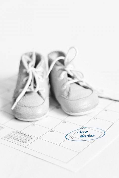 A pair of white baby size shoes on top of a white calendar with the words "Due Date" circled in blue pen.