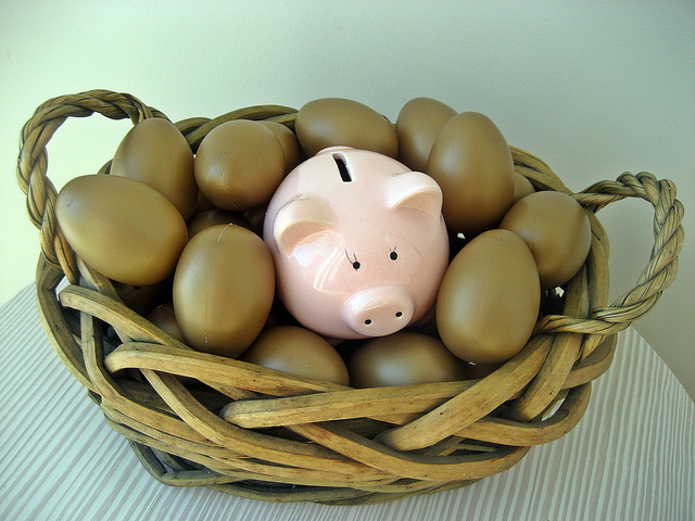 A small, pink ceramic piggy bank is nestled in the middle of a woven basket full of golden eggs.