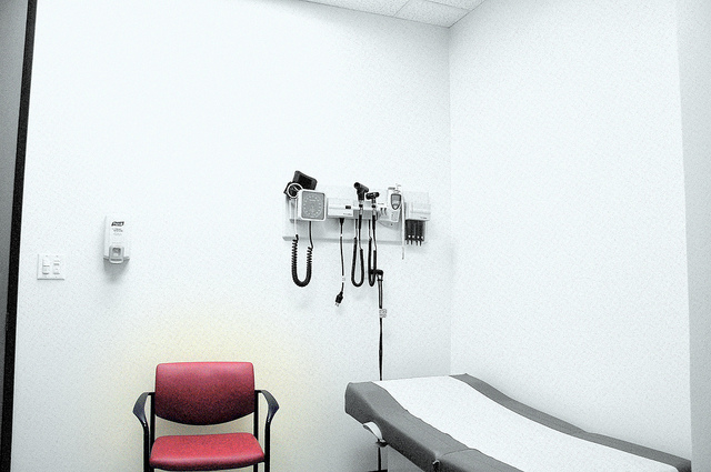 An empty doctors office, featuring a patient bed, a red chair, a hand sanitizer sipernser, and several instrumental tools on a wall.