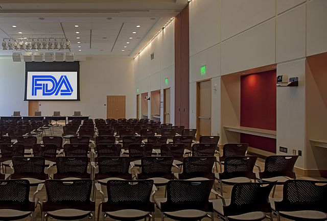 An empty room with several rows of chairs facing a stage with a screen that reads "FDA"