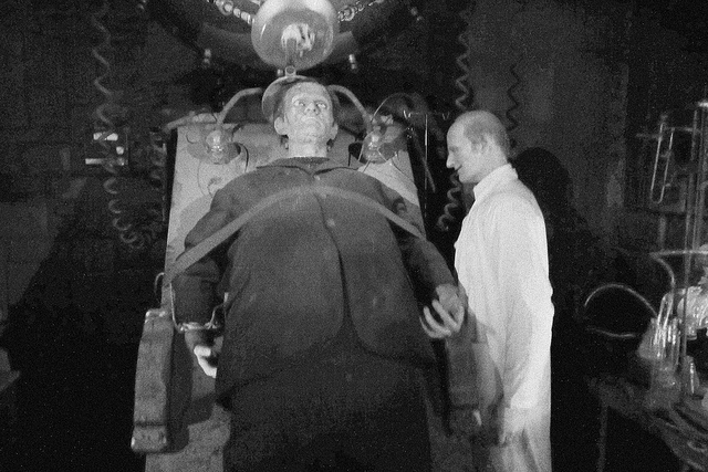 Black and white image of Dr. Frankenstein standing over his creation, a humanoid creature which lays strapped to a bed.