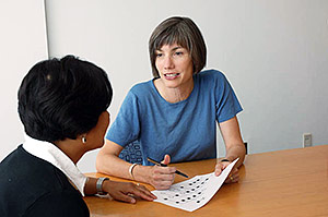 Two women sit together at a table. The woman to the right, a genetic counselor, holds a sheet of paper containing genetic information and speaks to the woman on the left.