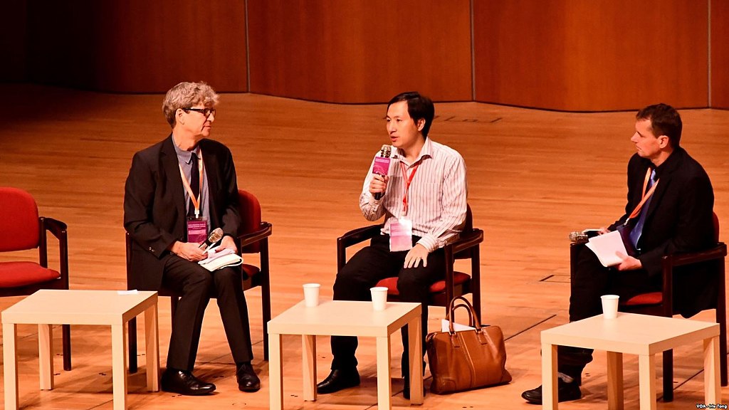 He Jiankui sitting and speaking into a microphone with a panel of speakers