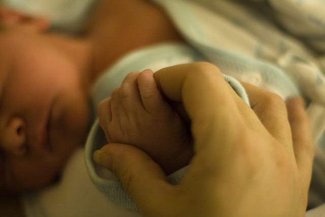 Close-up of a sleeping newborn baby whose hand is held by an adult with slender fingers.