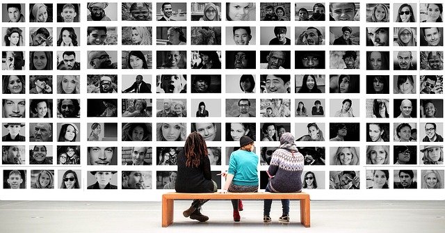 Three people sit with their backs turned as they look at a massive wall art exhibition of black and white portraits of people's faces.