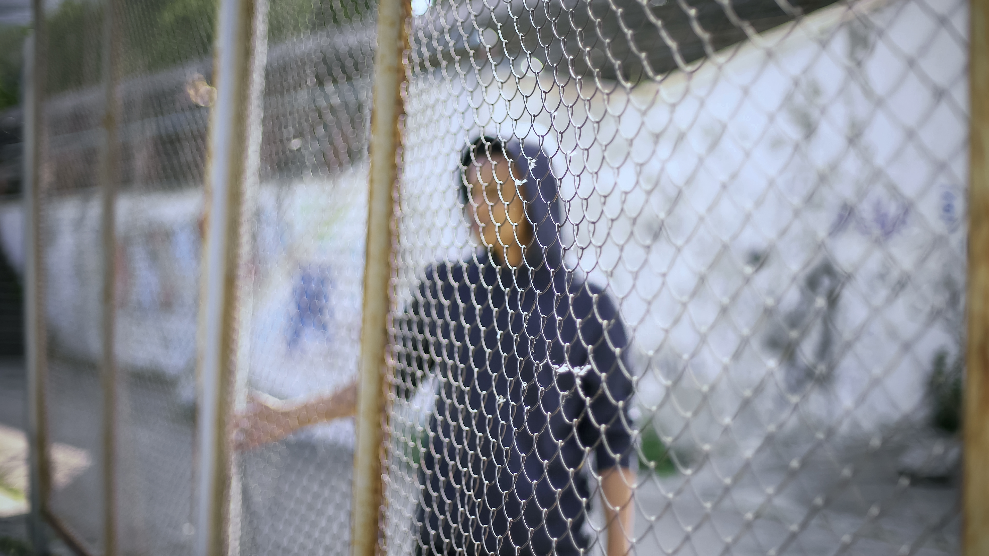 a boy in a blue hoodie stands behind a cyclone fence