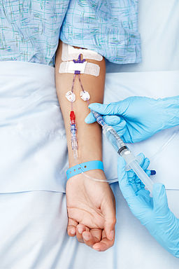 Bird's eye view of a patient's hand resting , with several wires hooked up, as a gloved hand inserts an IV needle.