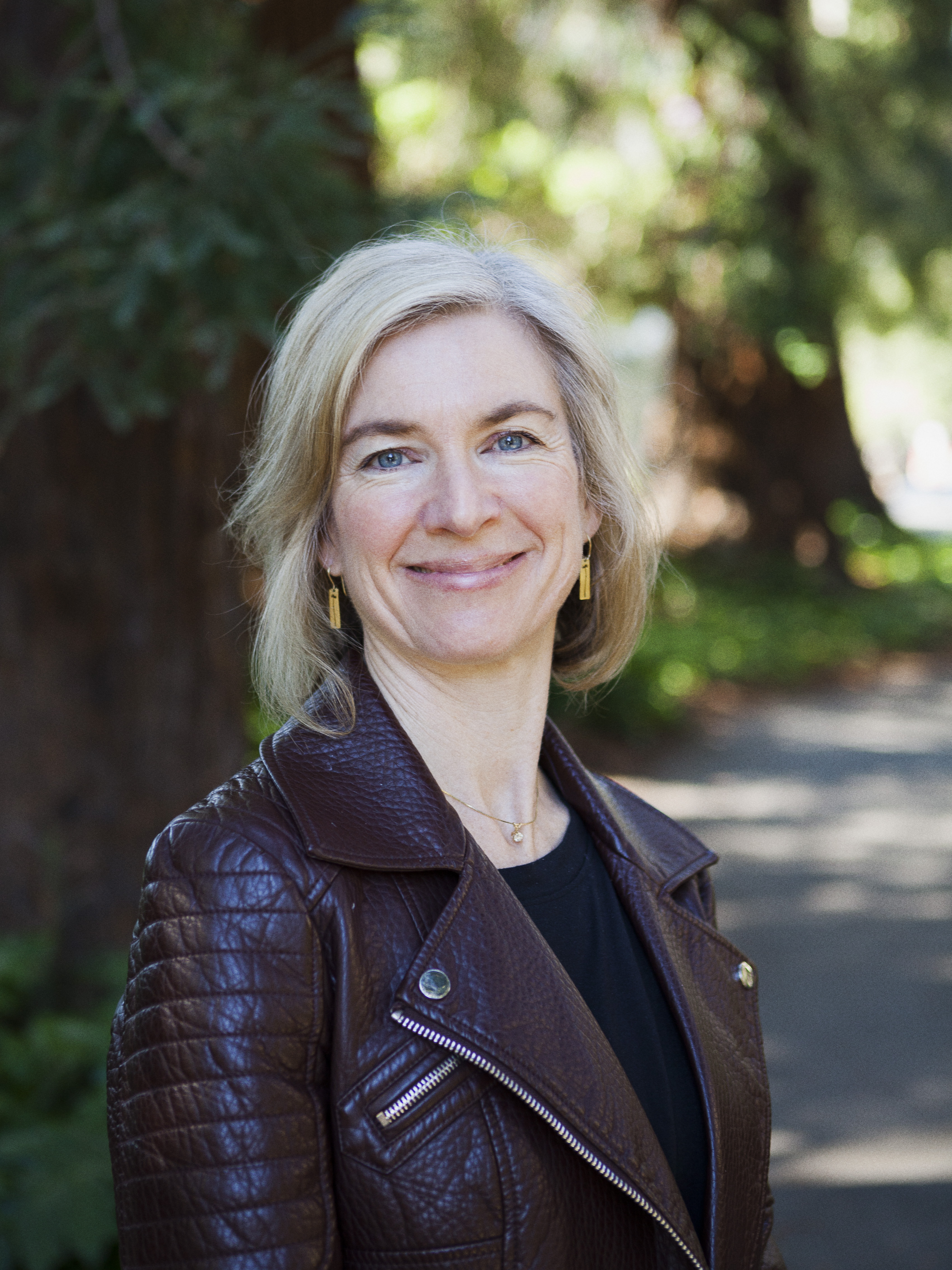 Portrait photo of Jennifer Doudna casually dressed and smiling in front of shaded trees.