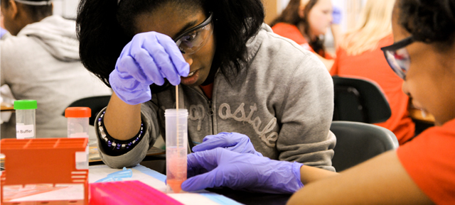 Two young women, with protective eye wear and gloved hands, use plastic lab materials to look at a test tube filled with a pink liquid