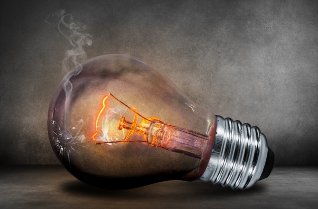 A cracked light bulb is tipped on its side,a weak current is visible causing a glow. The background has an eerie mix of black and white colors and shadow.