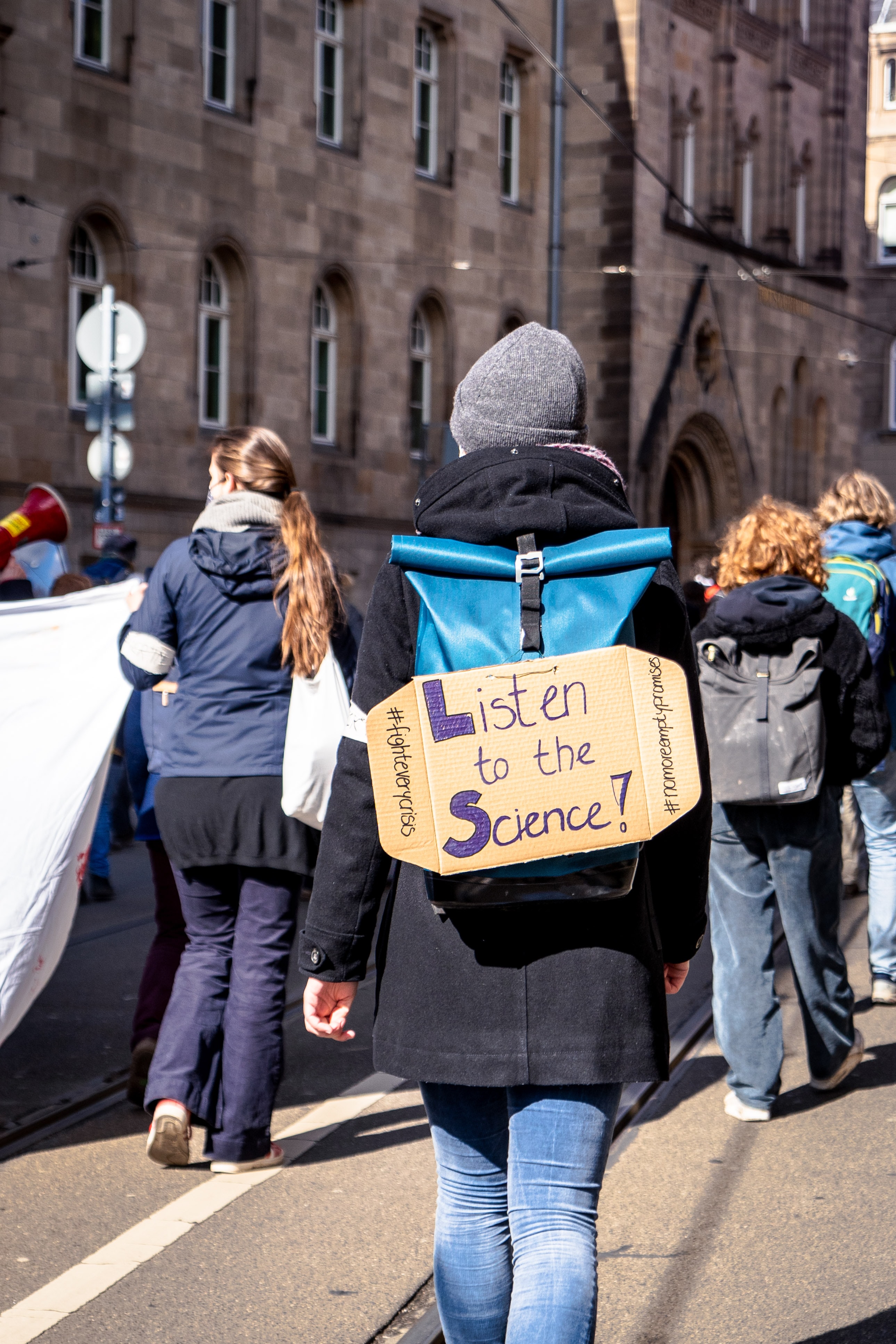 Woman protesting with a sign that says "Listen to the Science!" hanging on her backpack