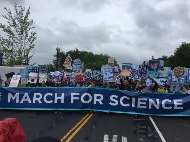 Several protesters hold signs behind a long banner stating "March for Science."