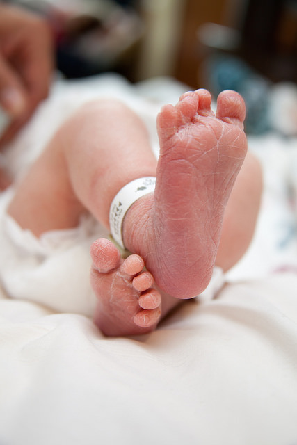 Close up of the feet of a newborn baby laying on a white bed in a hospital. The baby has a hospital tag around its right ankle.