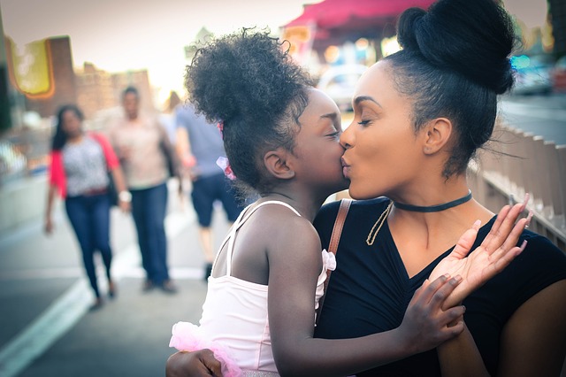 A black woman kisses a young black girl with joy and love expressed on their faces. In the background, there is a city back drop with other people walking on the sidewalk is featured, but out of focus and blurred.