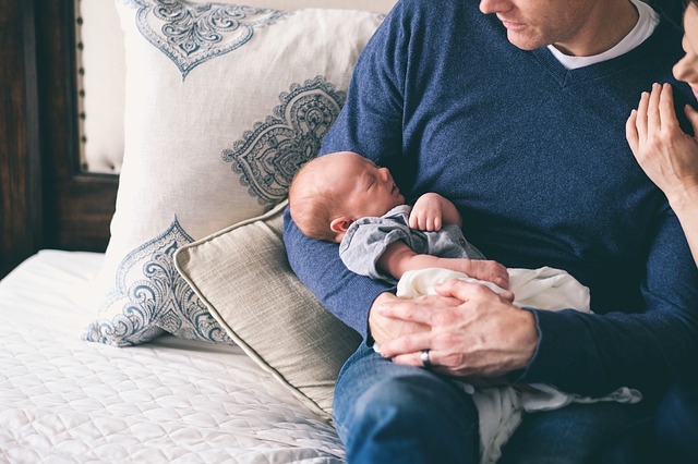 A photo showing a white man, seated on a couch, holding a newborn in his arms.