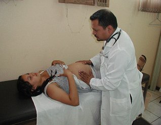 A male doctor examines a pregnant woman's exposed stomach as she lays does on a hospital bed.